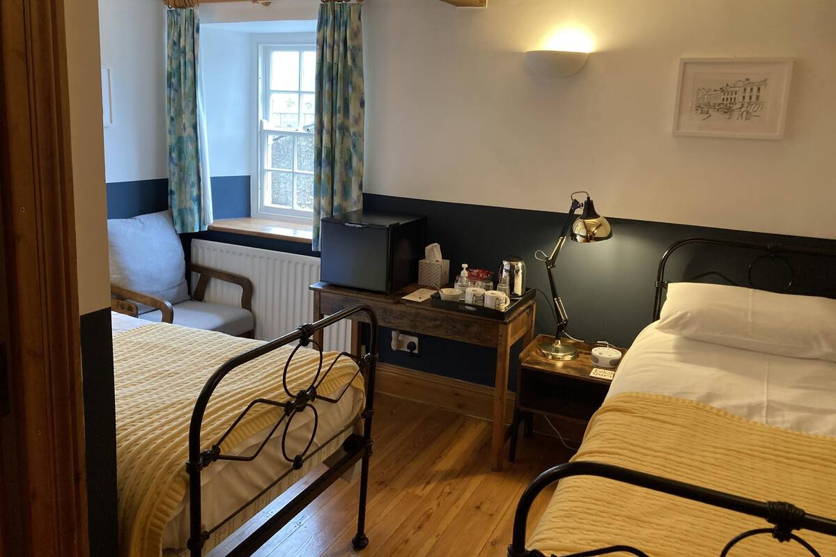 A room with two beds, a desk with a lamp, a chair, a radiator, and a window with curtains.