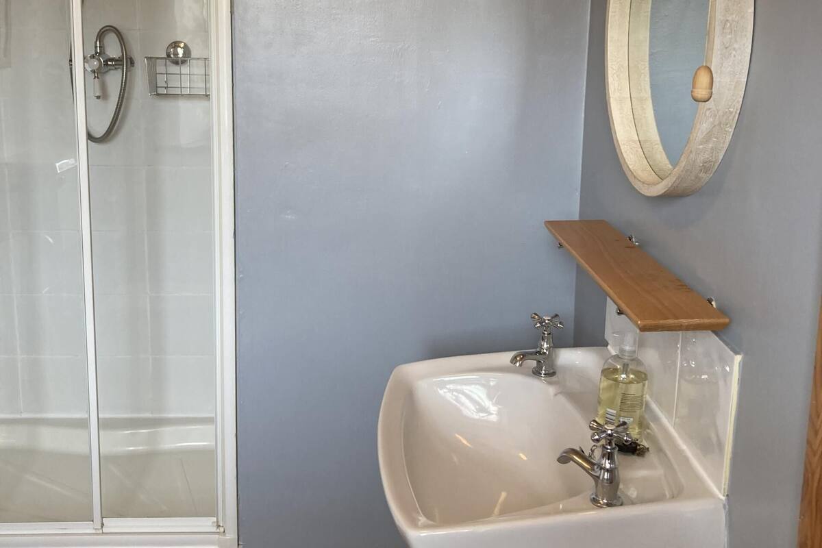 A bathroom with a glass shower enclosure, a white sink with a wooden shelf, and a round mirror on the wall.