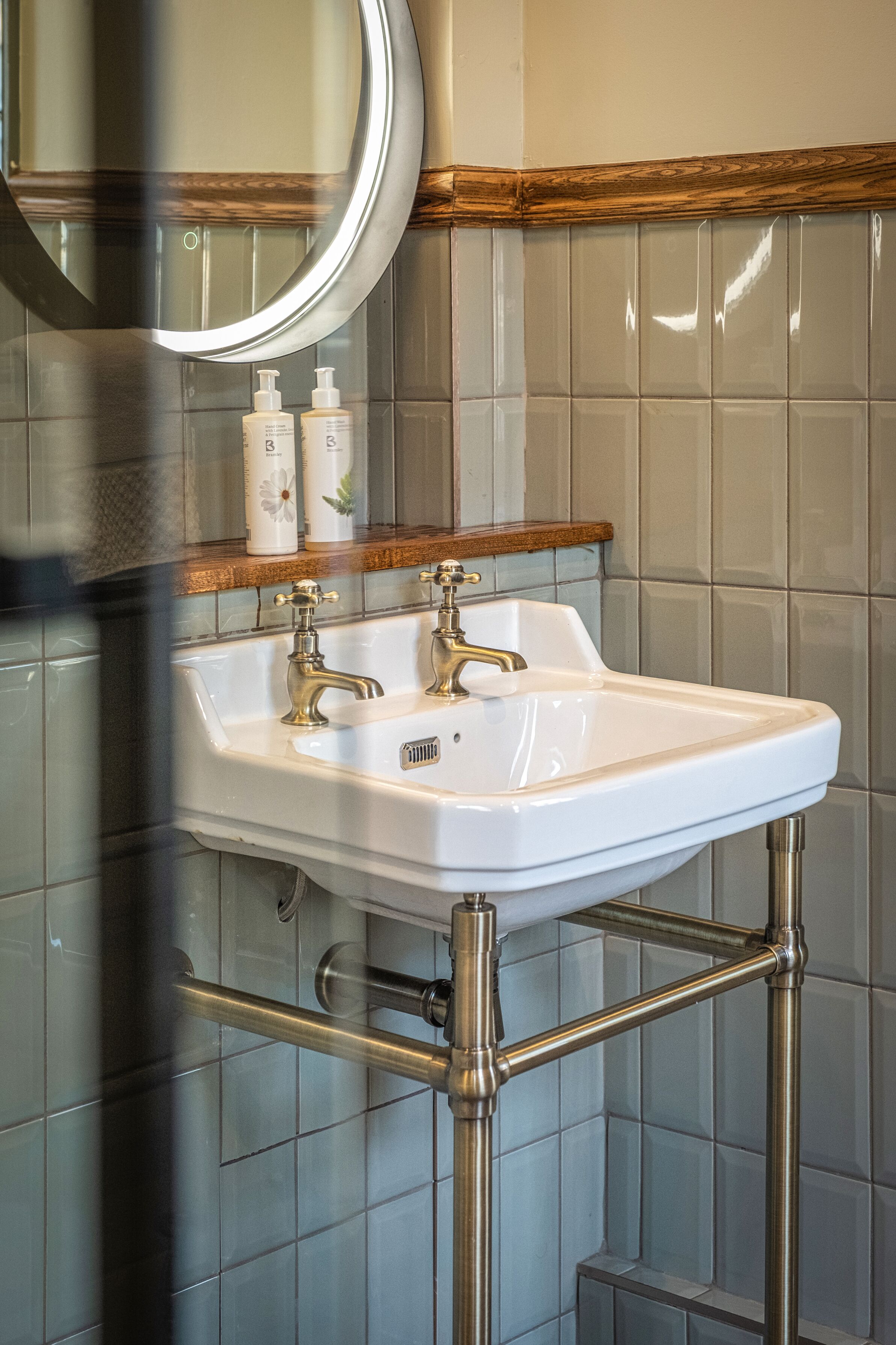 Ein Badezimmer mit einem weißen Waschbecken, zwei Wasserhähnen und einem Spiegel darüber. Auf dem Rand des Waschbeckens stehen zwei Flaschen.