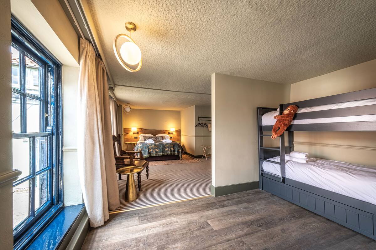 A bedroom with a bunk bed, a desk, and a chair. There is a window with curtains, a lamp, and a bed with a teddy bear on the top bunk.