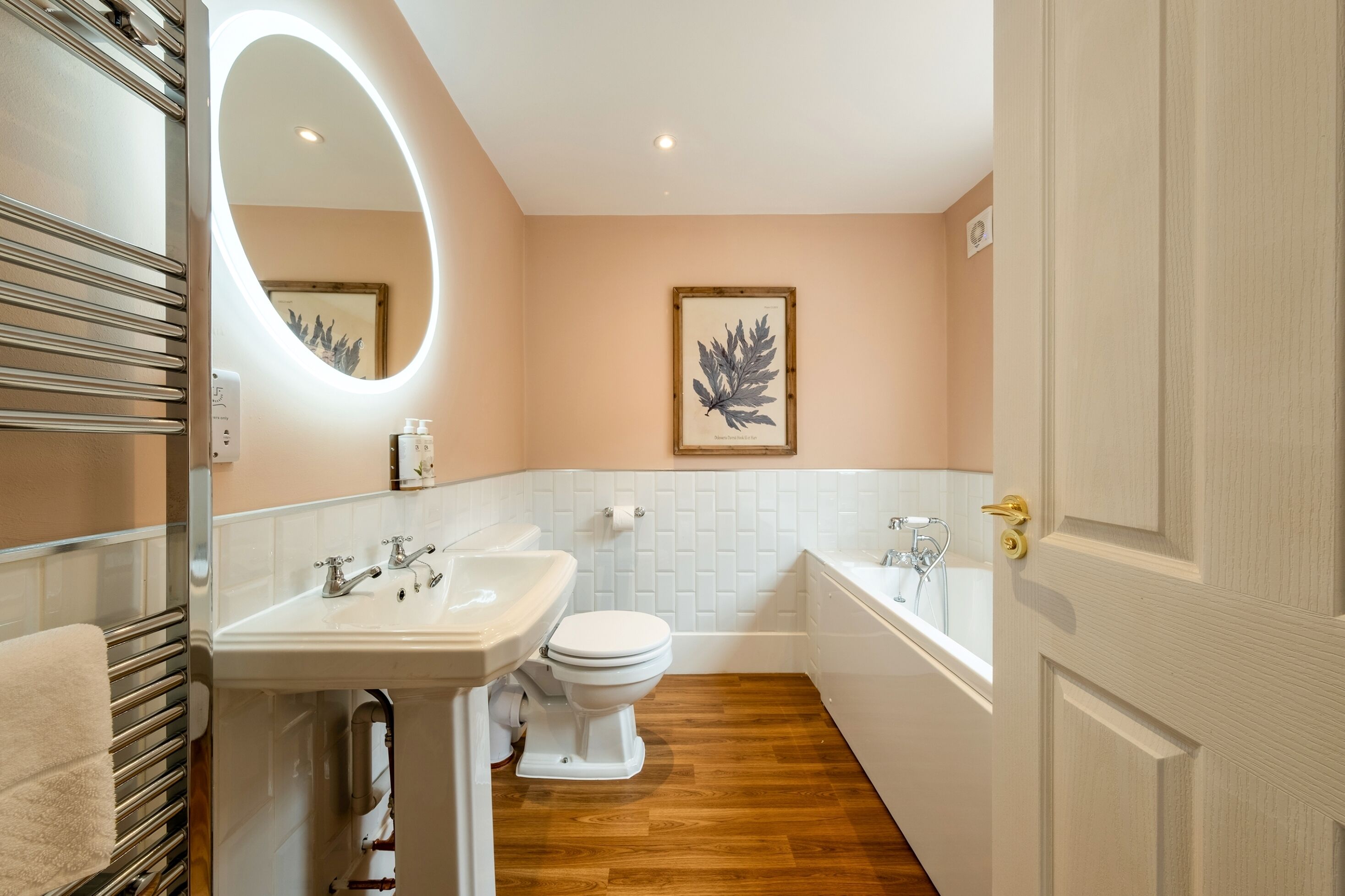 A modern bathroom with a wooden floor, white fixtures, and a large mirror with integrated lighting.