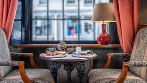 A dining area with a table set for breakfast, two patterned chairs, a window with a view of buildings, and a red lamp.
