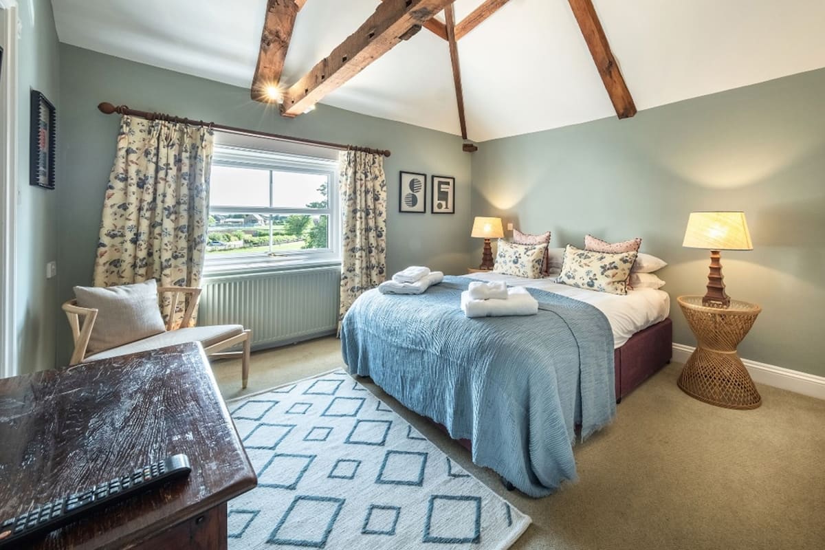 A bedroom with a bed, two bedside lamps, a window with curtains, a chair, and a desk.