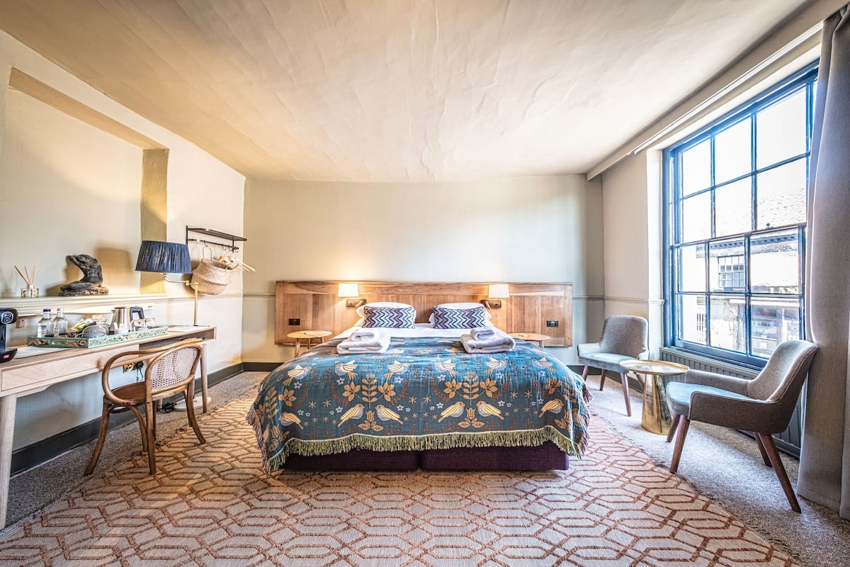 A hotel room with a large bed, a desk with a lamp, a chair, a small table, and a window with a view.