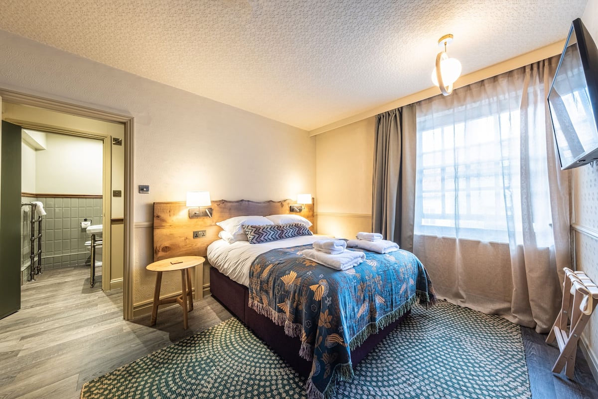 A hotel room with a bed, bedside lamps, a television, a window with curtains, and a bathroom visible through an open door.