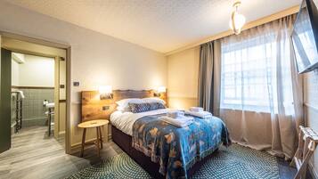 Ein Hotelzimmer mit einem Bett, Nachttischlampen, einem Fernseher, einem Fenster mit Vorhängen und einem durchgegangene Tür sichtbaren Badezimmer.