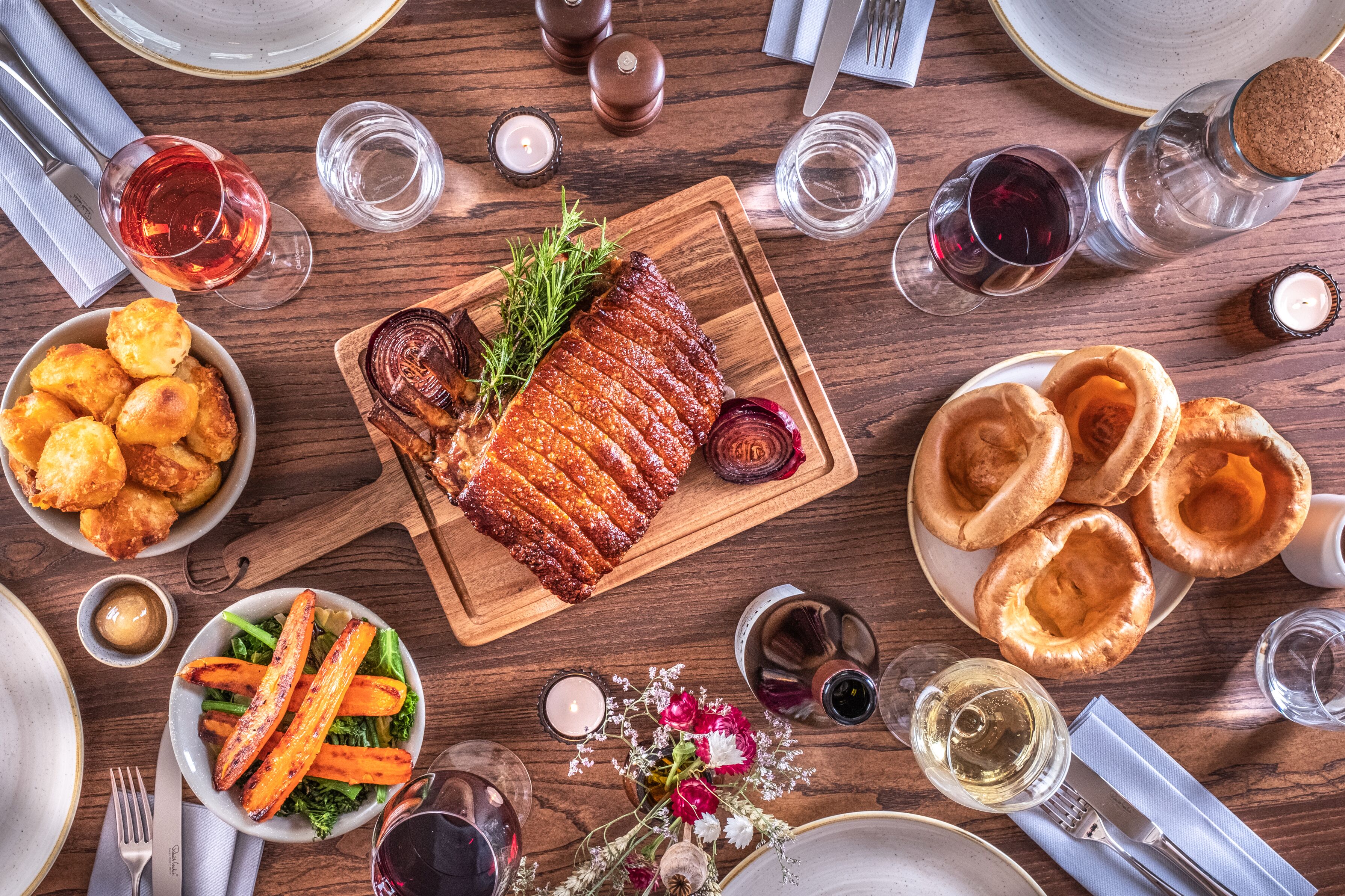 A dining table with a variety of dishes, including roasted meat, Yorkshire puddings, and roasted vegetables, surrounded by glasses of wine.