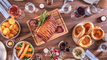 A dining table with a variety of dishes, including roasted meat, Yorkshire puddings, and roasted vegetables, surrounded by glasses of wine.
