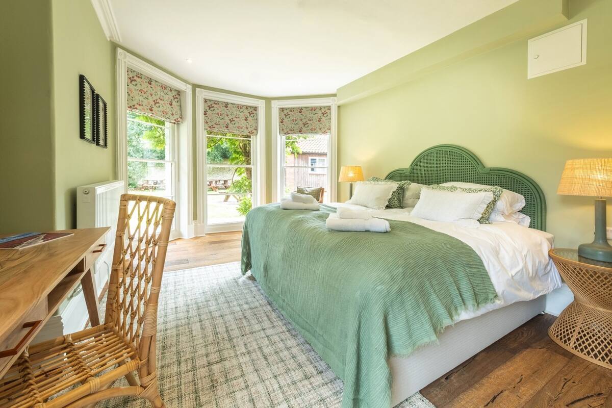 A bedroom with a bed, a wooden desk, a wicker chair, and a view of the outdoors.