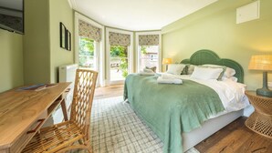 A bedroom with a bed, a wooden desk, a wicker chair, and a view of the outdoors.