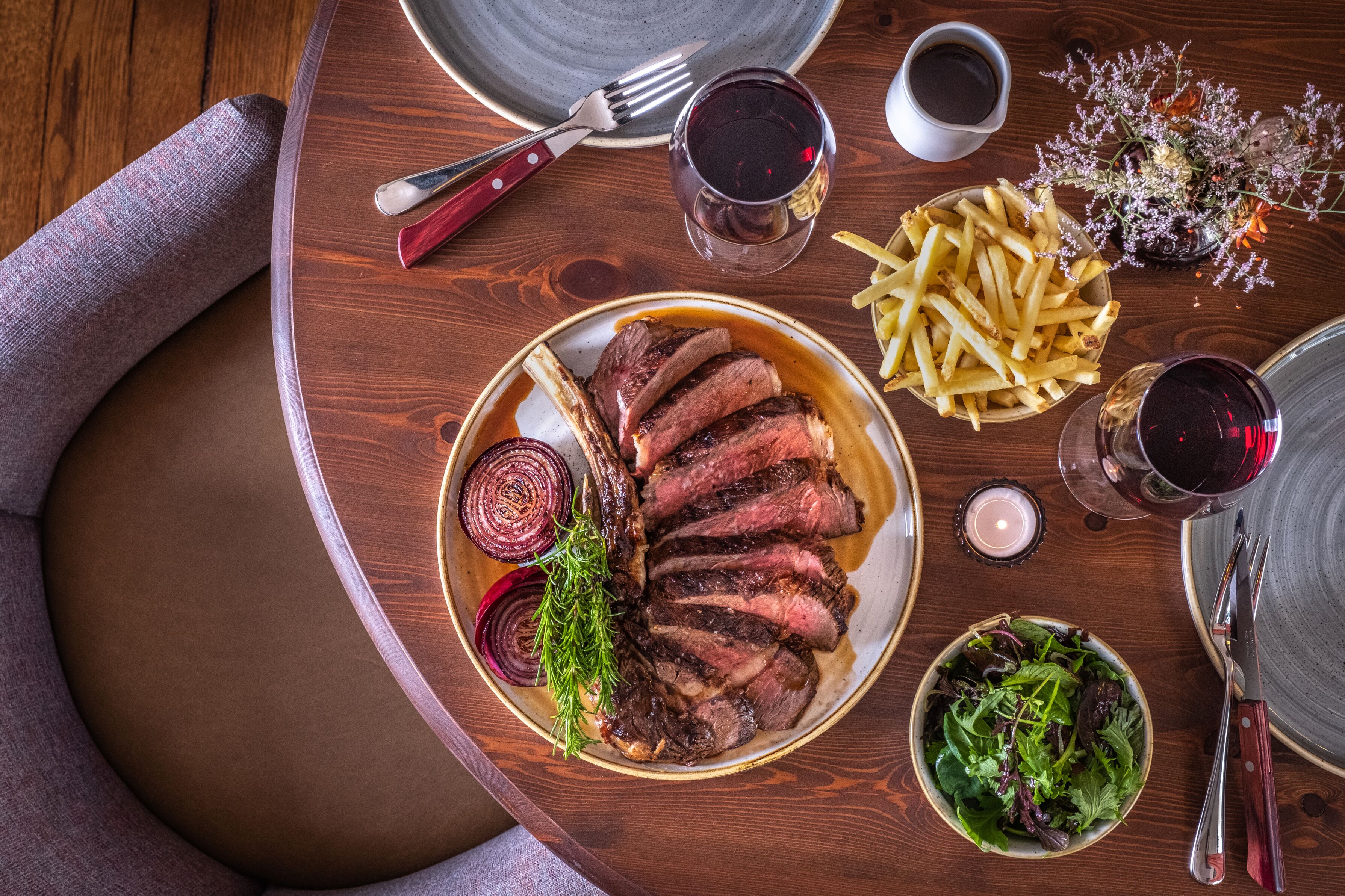 Una mesa de comedor con un plato de carne en rodajas, un bol de papas fritas, una ensalada pequeña y dos copas de vino tinto.