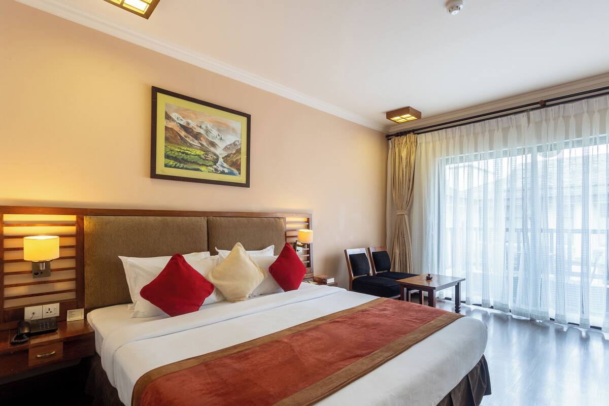 Una habitación de hotel con una cama grande, una mesita de noche con lámpara, un escritorio con dos sillas, una ventana con cortinas translúcidas y un cuadro en la pared.