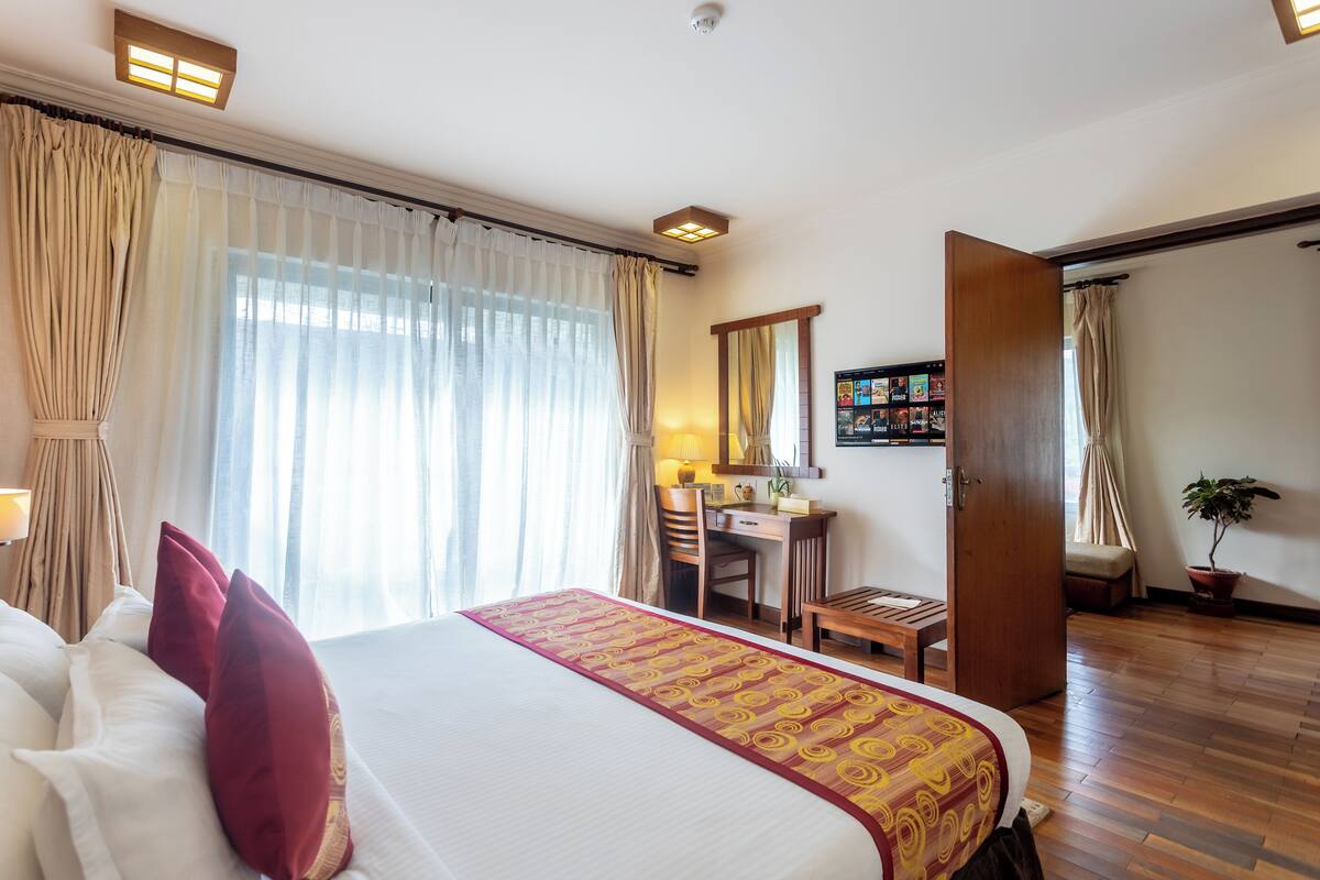 Una habitación de hotel con una cama grande, un escritorio, una silla, una mesita, una planta en maceta y una puerta que da a otra habitación.