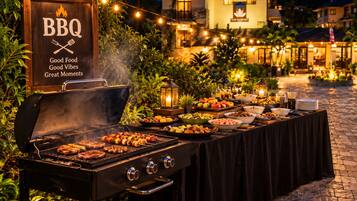 An outdoor BBQ setup with a grill, skewers, and a variety of dishes on a table, surrounded by string lights and a building in the background.