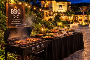 An outdoor BBQ setup with a grill, skewers, and a variety of dishes on a table, surrounded by string lights and a building in the background. - Atithi Resort & Spa (Pokhara)
