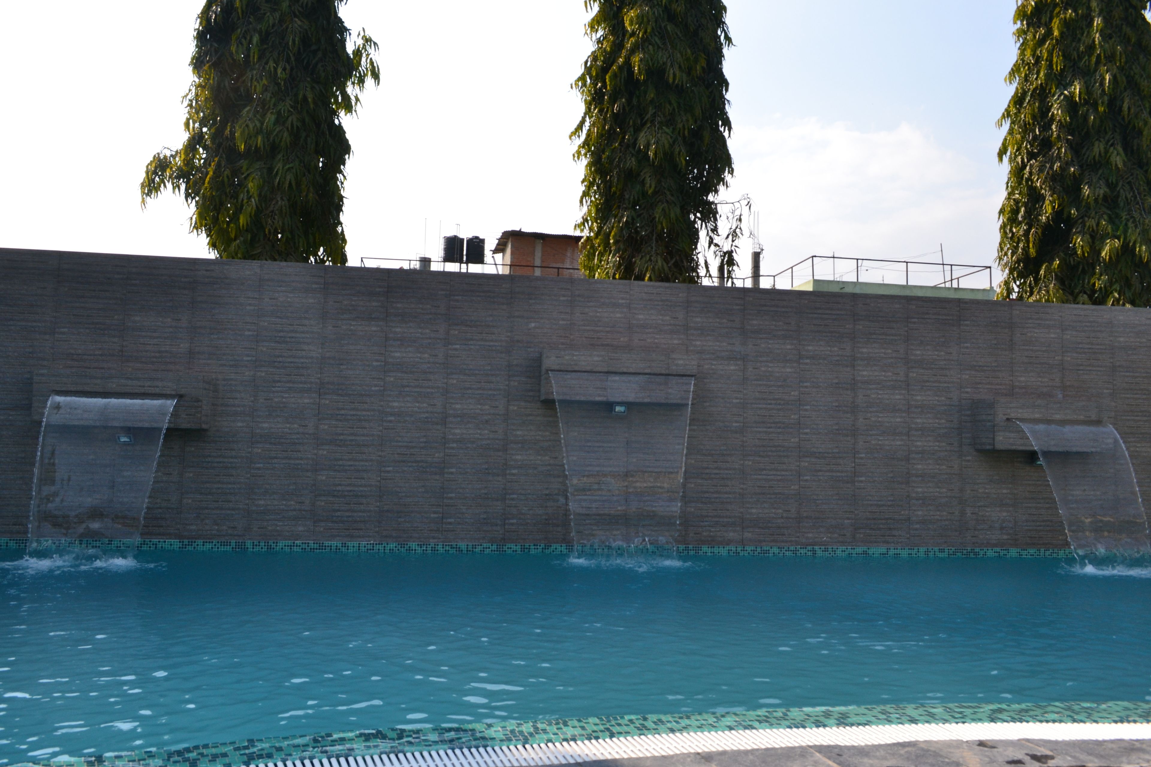 Una piscina con borde de azulejos, una pared de concreto con tres desagües, y altos árboles al fondo.