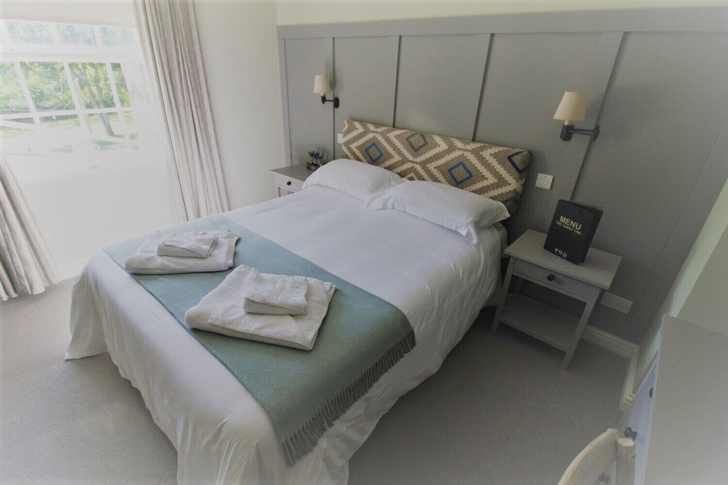 A neatly made bed with a pillow and two folded towels, a bedside table with a menu, and a window with curtains.