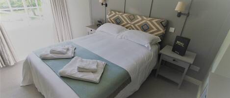 Una cama bien tendida con una almohada y dos toallas dobladas, una mesita de noche con un menú, y una ventana con cortinas.