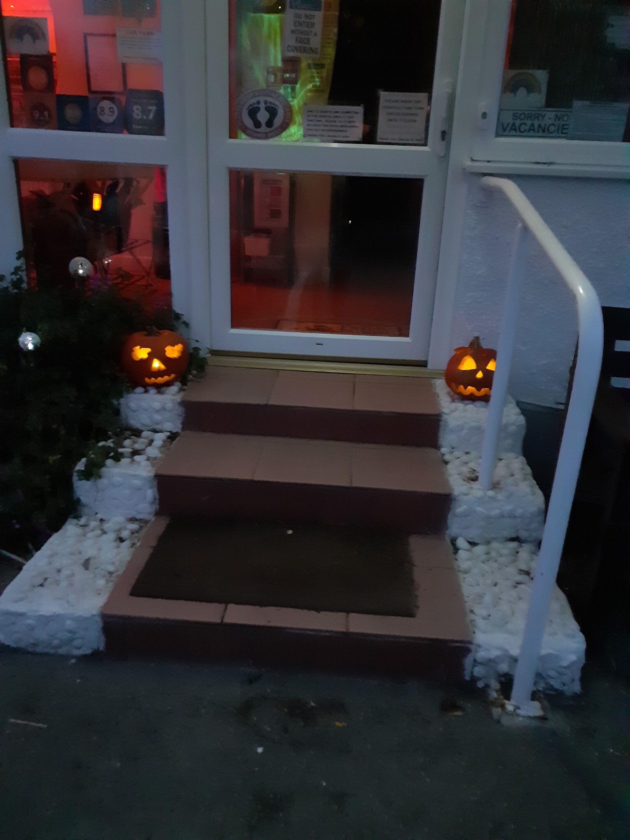 A set of outdoor steps leading to a door with a 'No Vacancy' sign, surrounded by white pebbles and two illuminated jack-o'-lanterns.