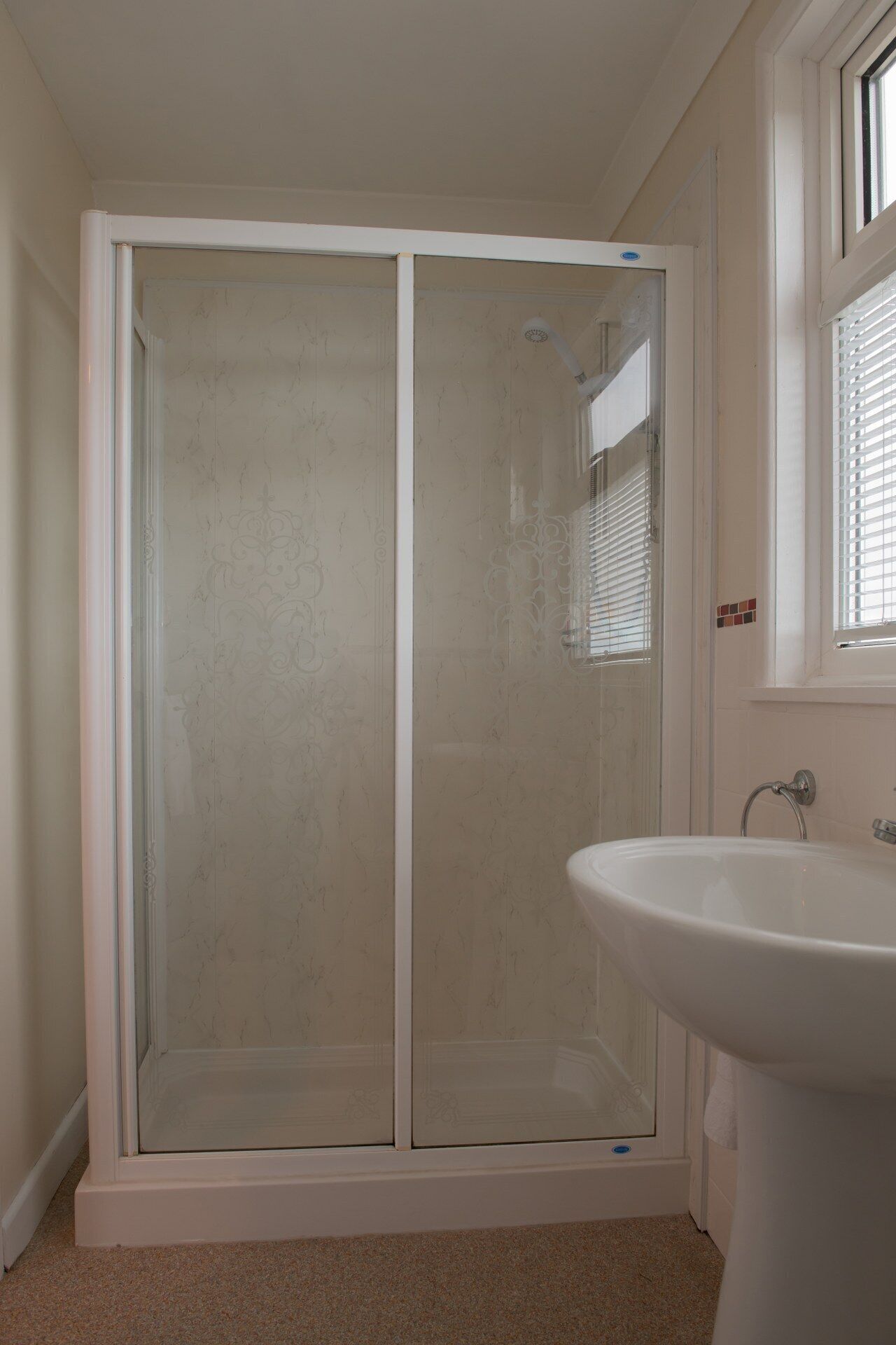 Une salle de bain avec une douche à parois de verre, un lavabo blanc et une fenêtre avec des stores.