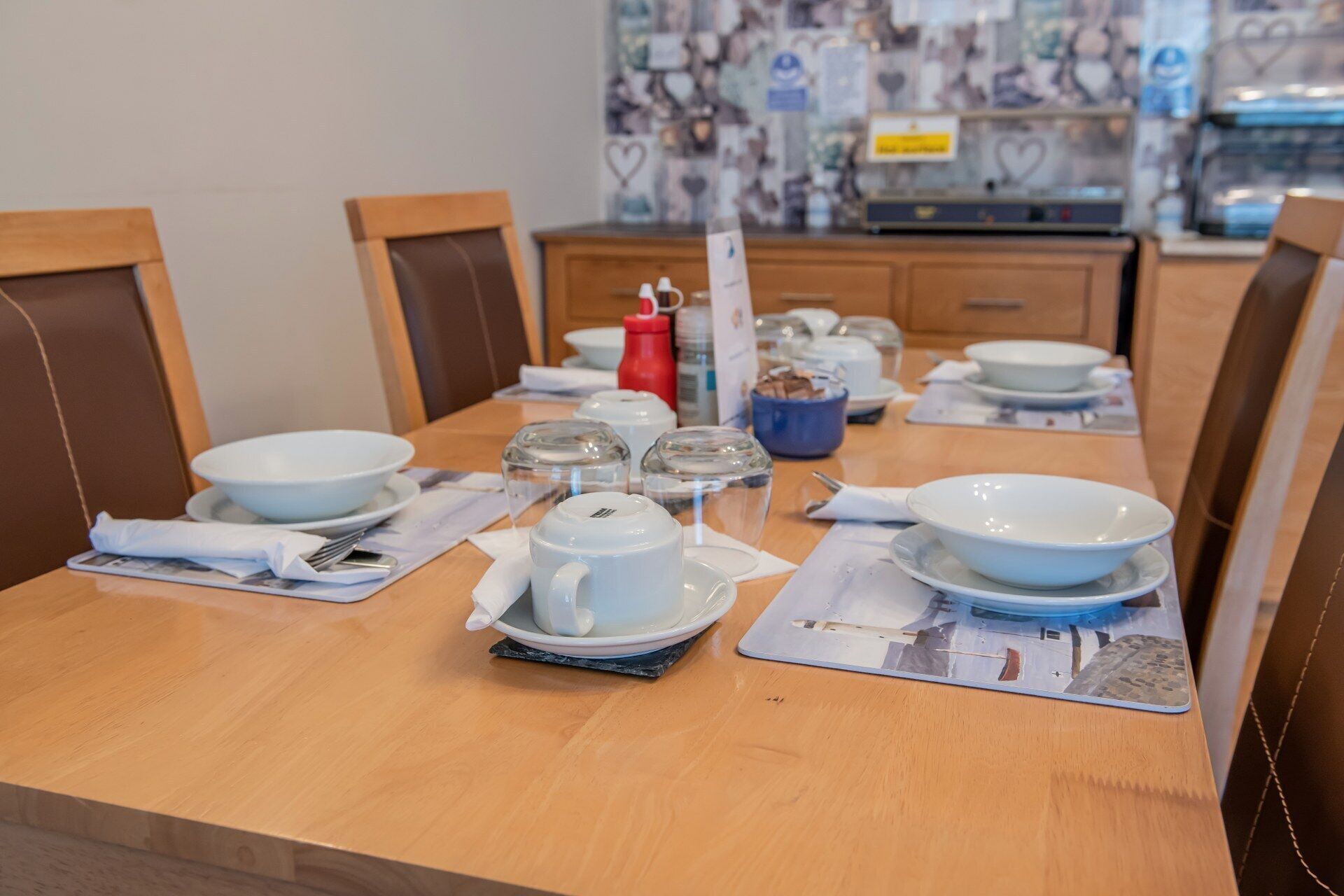 A dining table set with white plates, bowls, and a teapot, with chairs around it.
