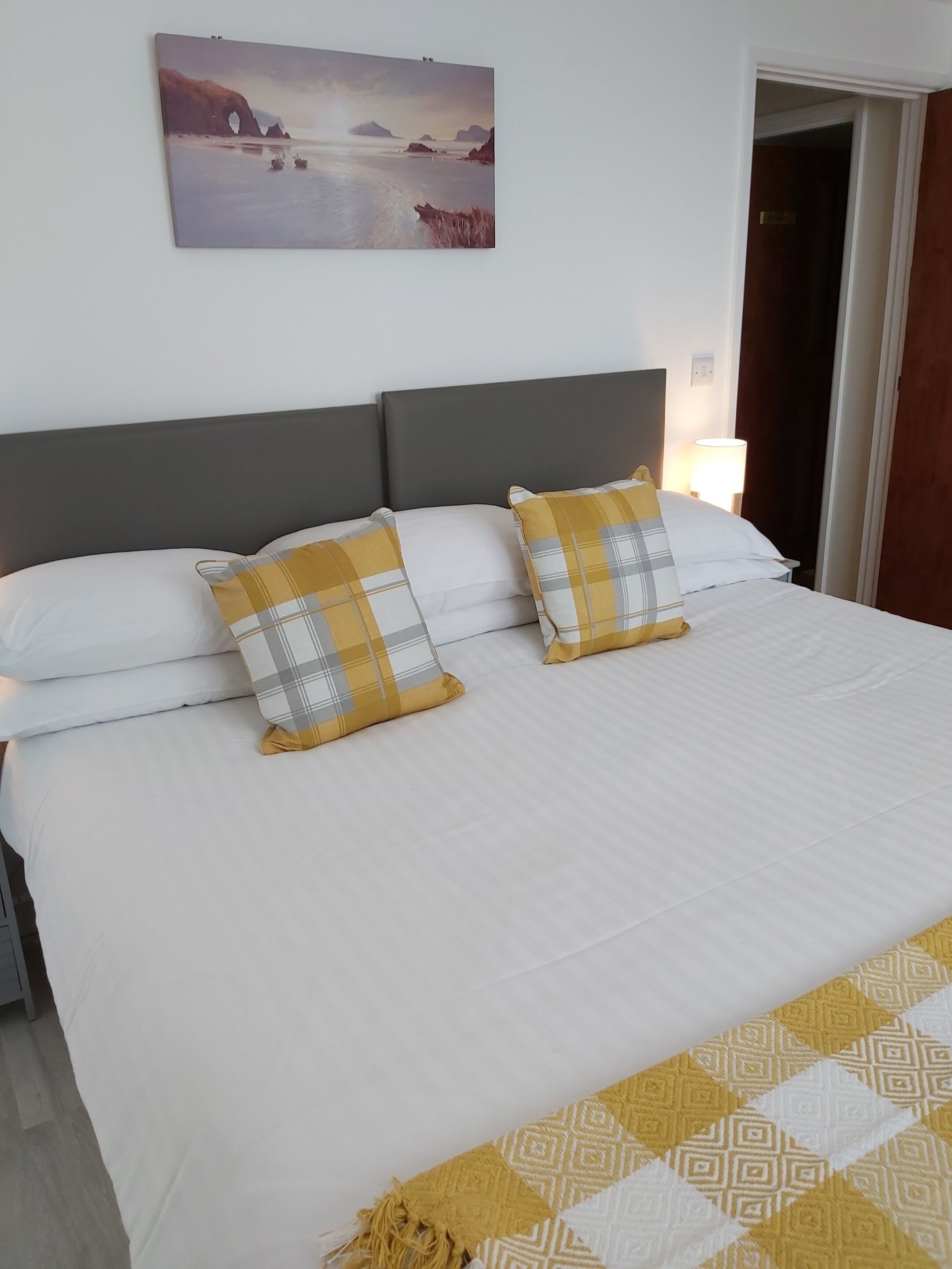 A neatly made bed with white and yellow bedding, a gray headboard, and a painting of a coastal scene above it.