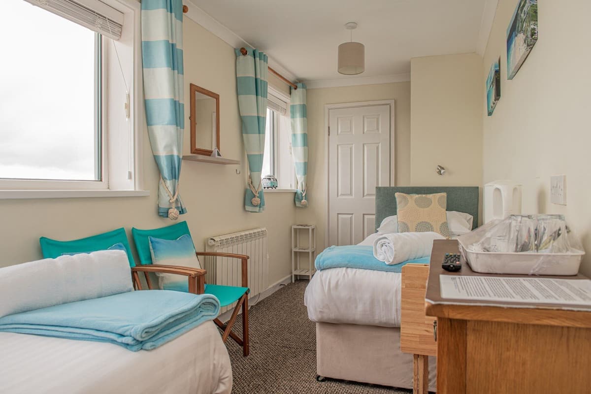 A hotel room with two beds, a desk, a chair, a radiator, a small table, and a window with curtains.