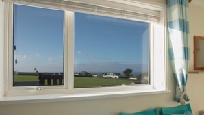A room with a large window overlooking a grassy field and buildings. There is a white window frame, a blue curtain, and a wooden chair with a cushion. - Trevarrian Lodge (Newquay)