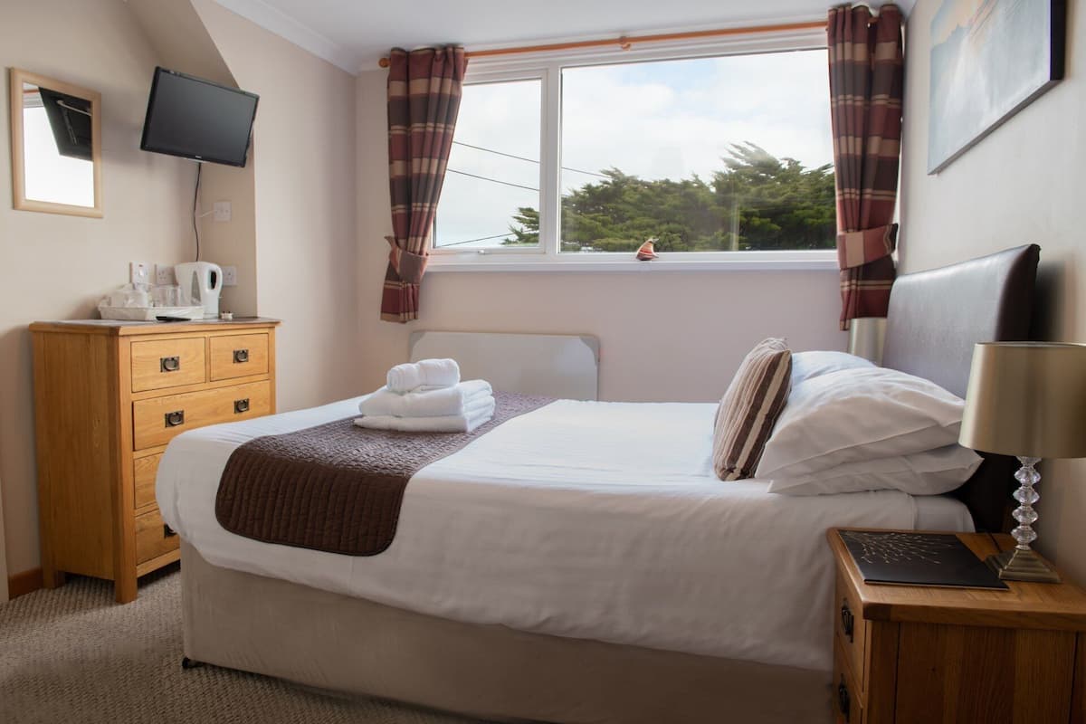 A neatly made bed with white linens and a brown throw, a wooden nightstand with a lamp, a wooden dresser with a flat-screen TV, and a window with plaid curtains.