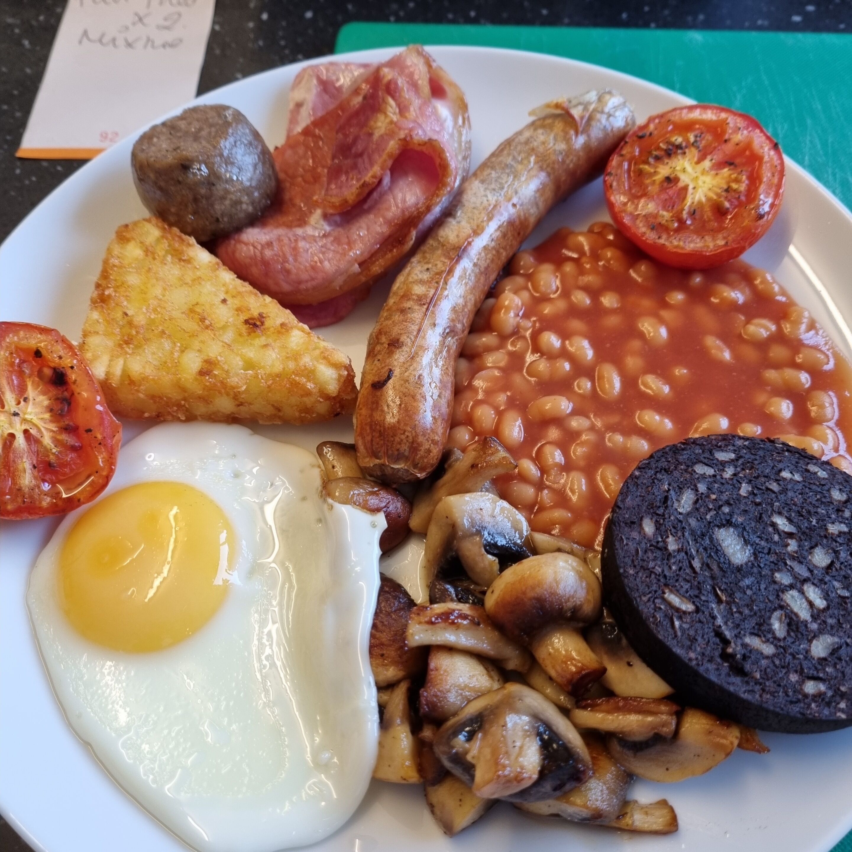 A plate of breakfast food including eggs, bacon, sausages, mushrooms, baked beans, and a slice of black pudding.
