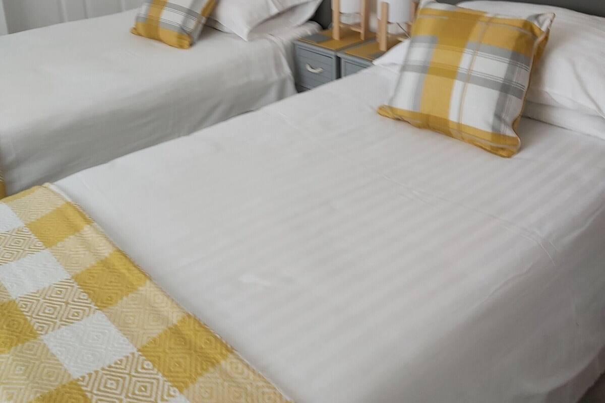 A hotel room with two single beds, each with a white and yellow checkered bedspread, a gray headboard, and a matching pillow. There is a nightstand with two white lamps and a small table with a yellow and white checkered tablecloth. A painting of a landscape is visible on the wall above the beds.