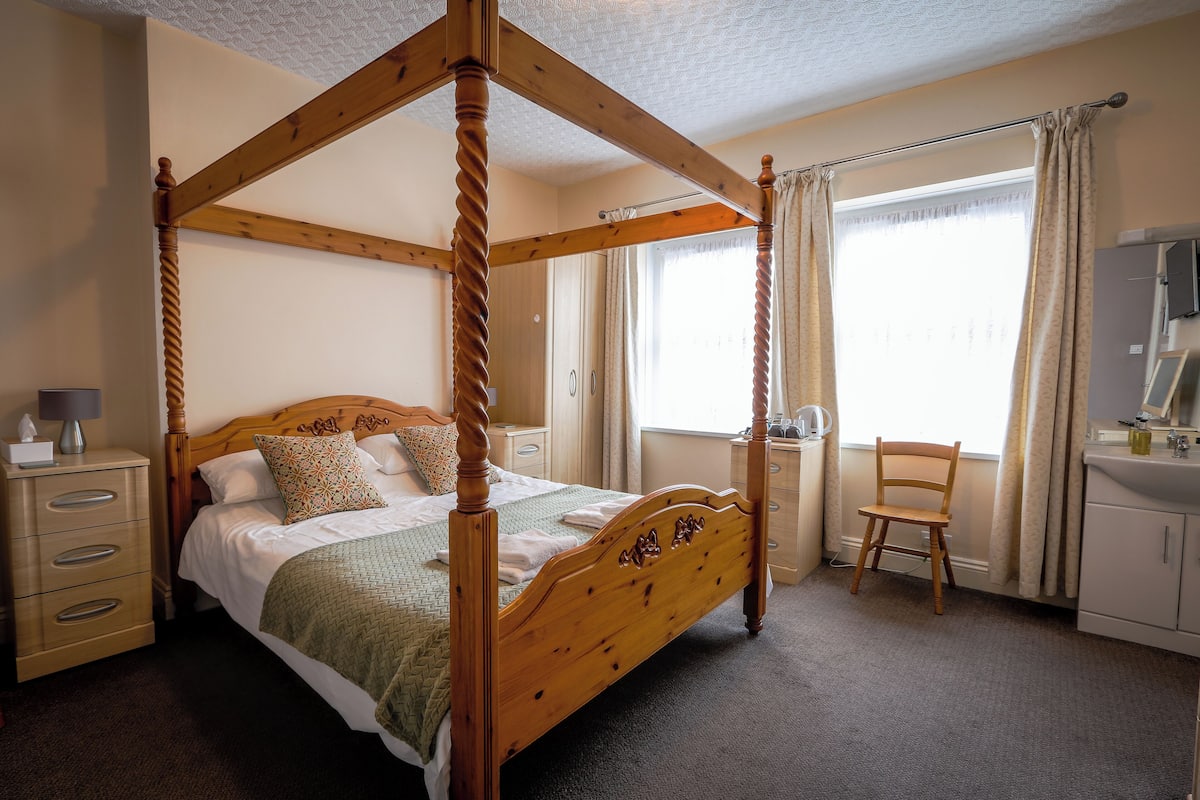 Una cama con dosel, un cabecero de madera, una mesita de noche con lámpara, una silla y una ventana con cortinas.