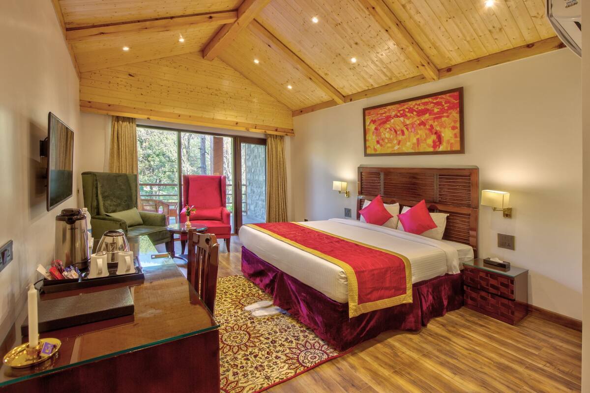 Une chambre d’hôtel avec un plafond en bois, un lit avec une literie rouge et blanche, un fauteuil vert, un fauteuil rouge, une table basse avec un plateau, et une grande fenêtre avec des rideaux.