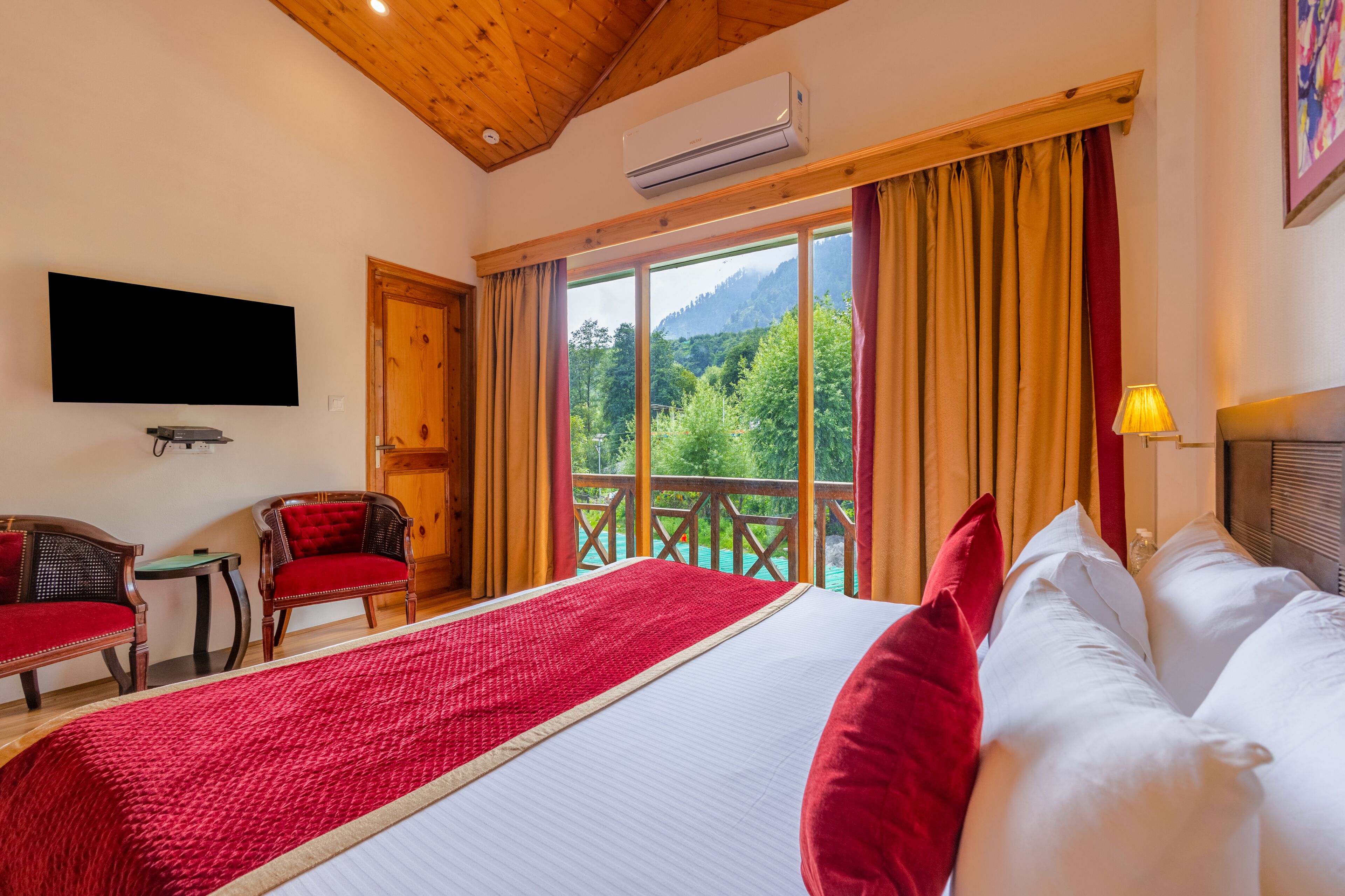 A hotel room with a large bed, a TV, a chair, and a balcony with a mountain view.