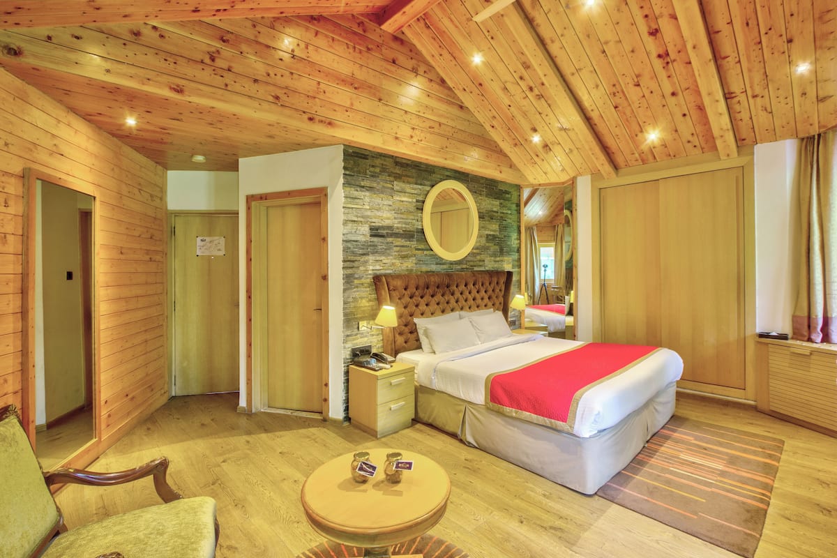Une pièce chaleureuse avec un plafond en bois, un lit recouvert d’une couverture rouge, un miroir rond et une petite table sur laquelle reposent deux objets.