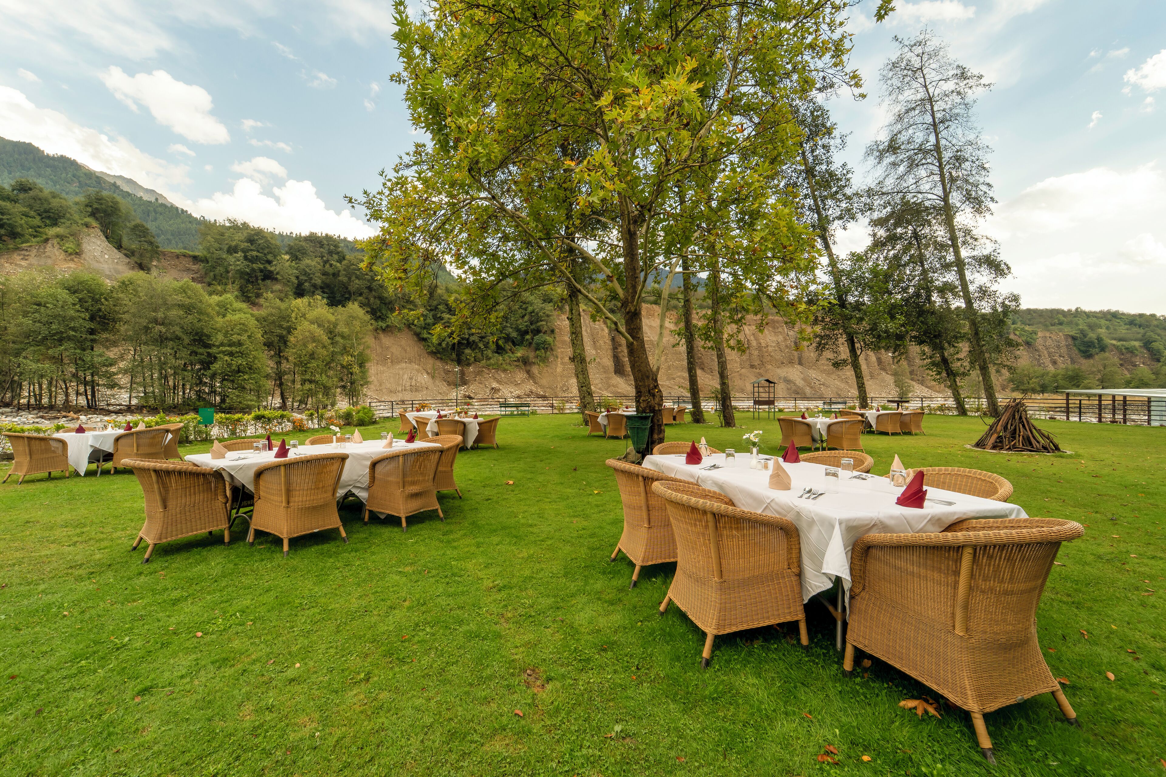 An outdoor dining area with wicker tables and chairs, surrounded by greenery and hills.
