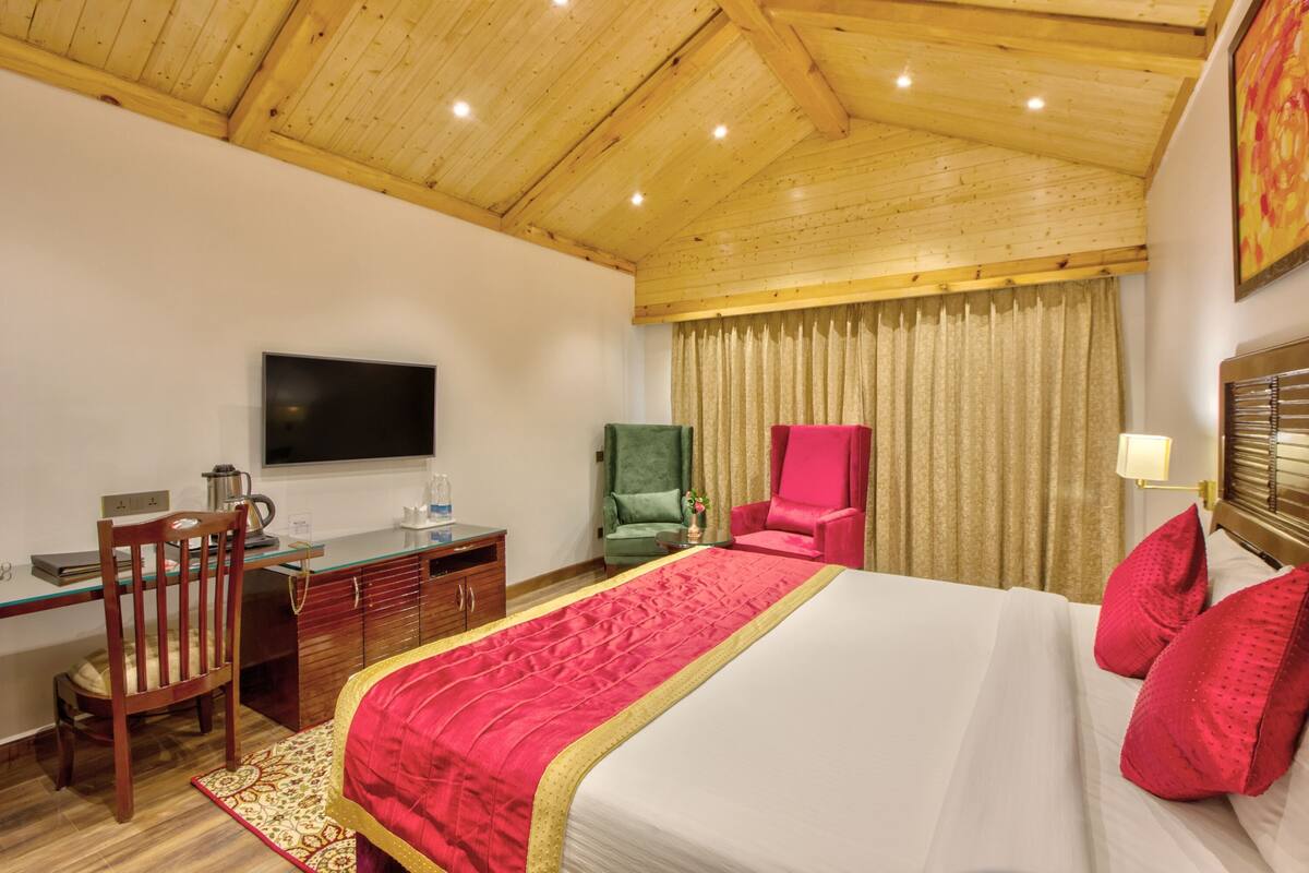 Une chambre d’hôtel avec un plafond en bois, un lit avec une literie rouge, un bureau avec une chaise, une télévision et une fenêtre avec des rideaux.