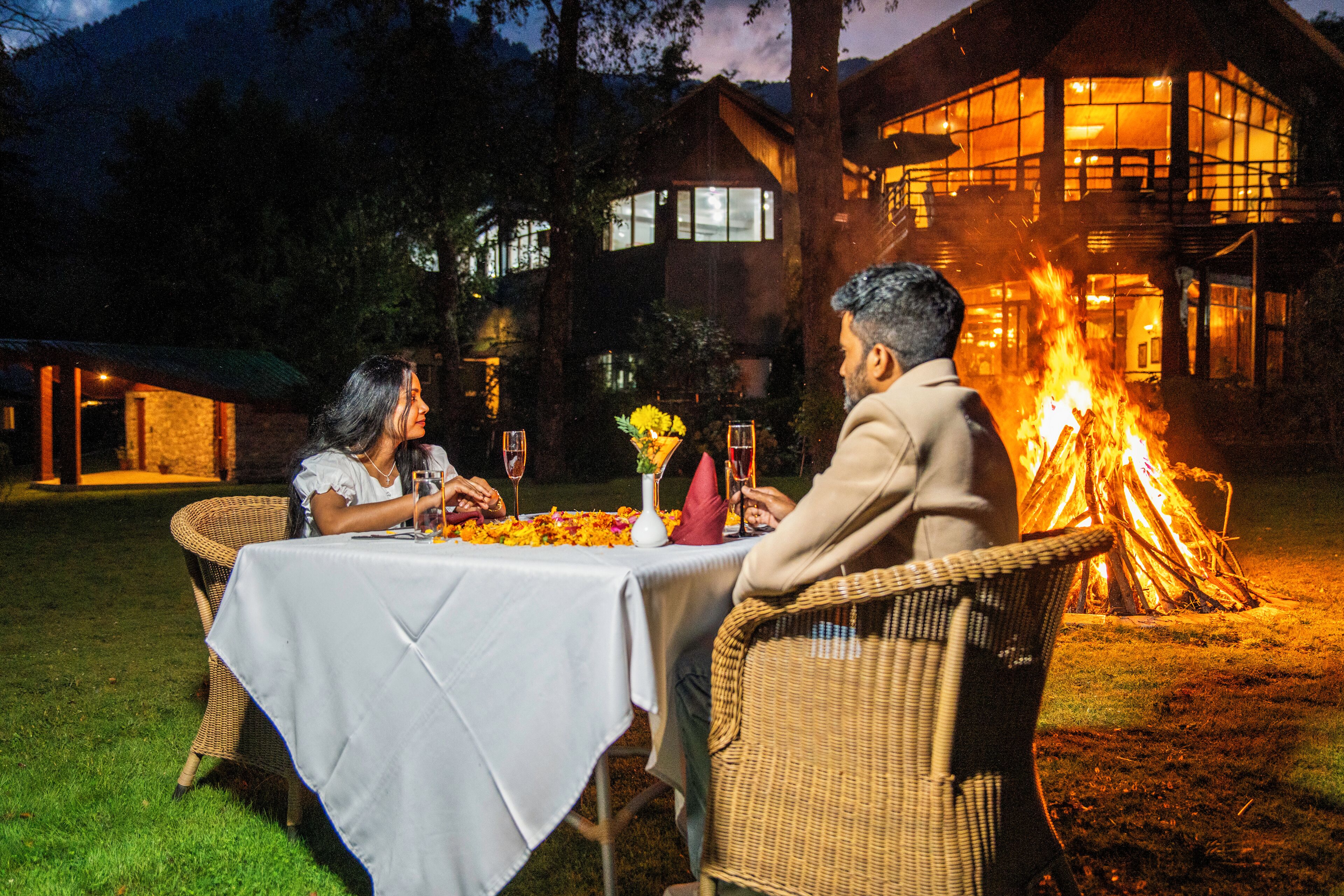 A couple dining at an outdoor table with a fire in the background.