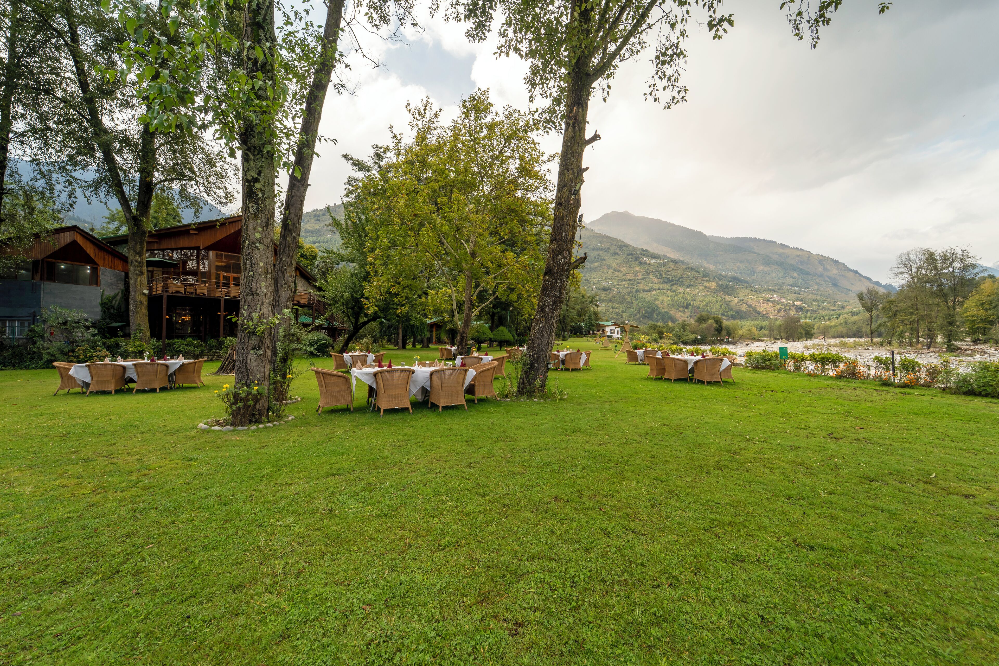 An outdoor dining area with tables and chairs, surrounded by trees and a mountainous backdrop.