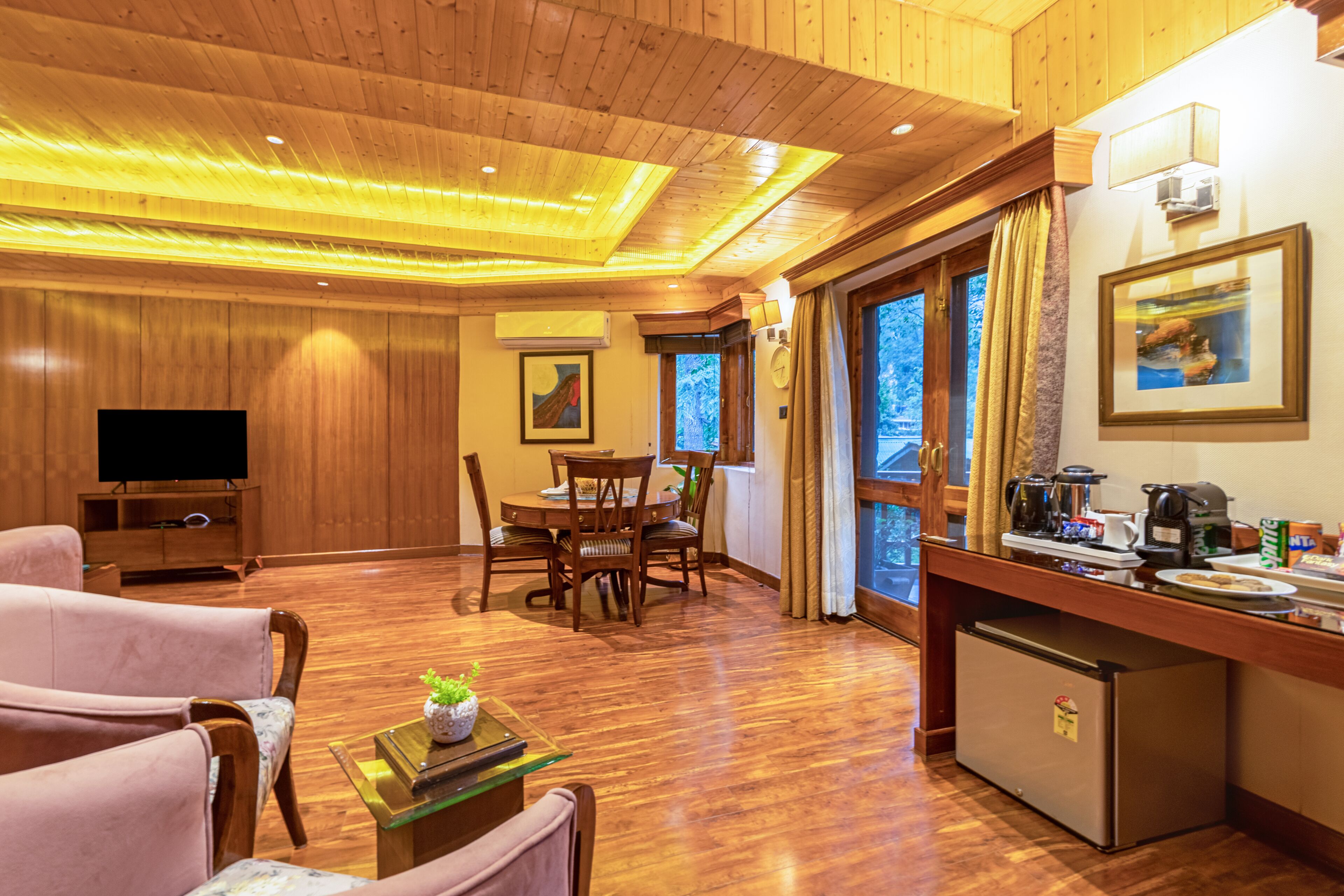 A hotel room with a wooden floor, a dining area with a table and chairs, a television, and a kitchenette with a refrigerator.