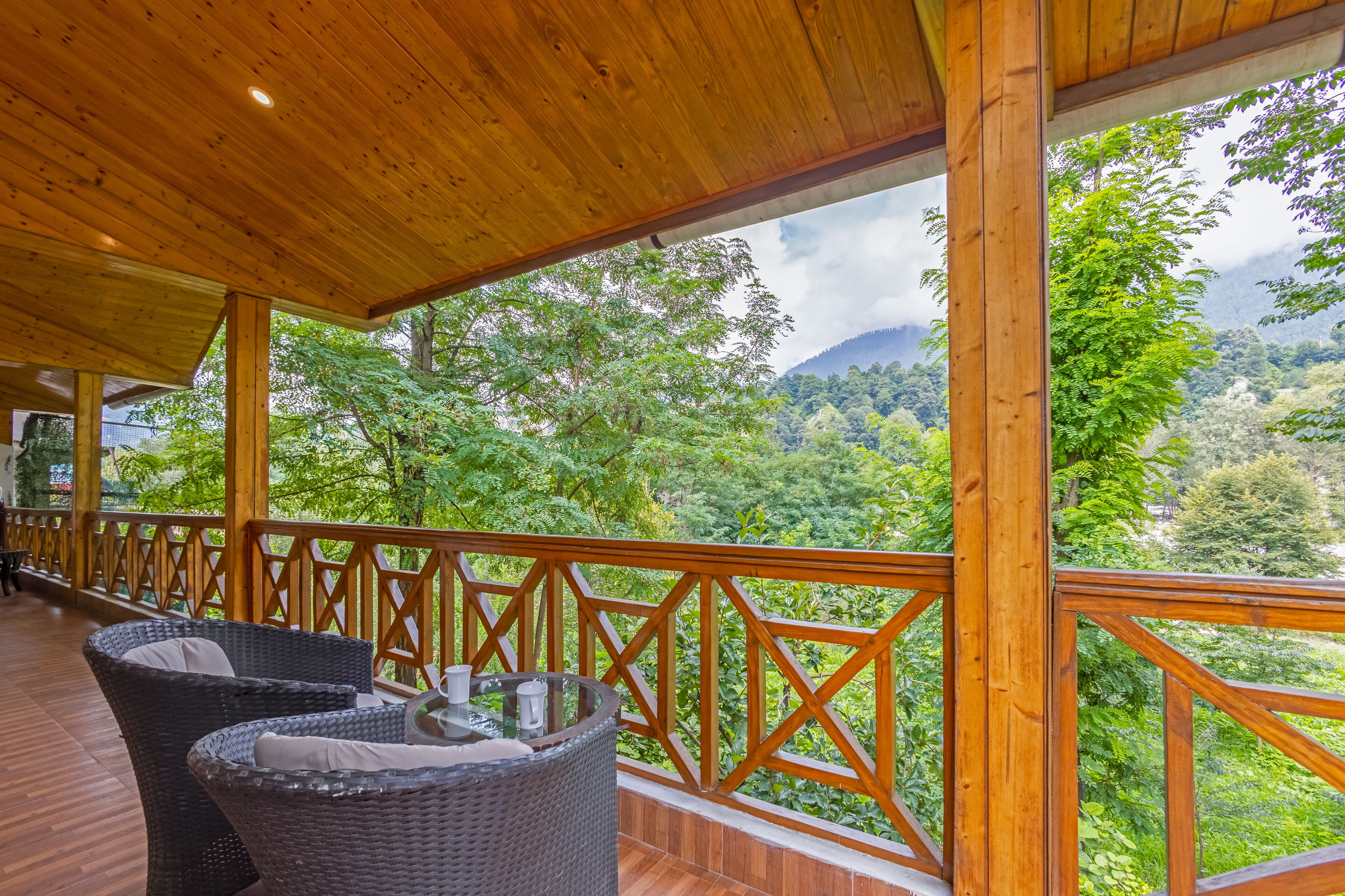Un espacio al aire libre techado con mobiliario de mimbre y barandilla de madera con vistas a un valle boscoso.