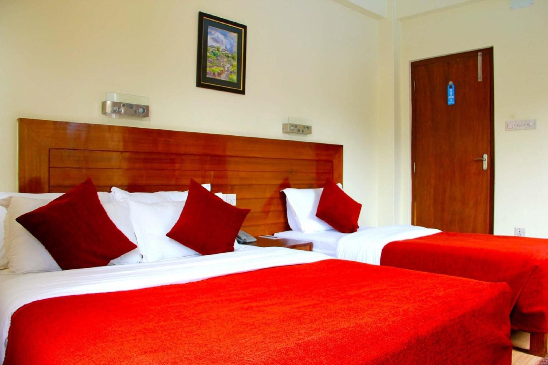 A hotel room with two beds, each with red bedding and pillows. There is a wooden headboard, a framed picture on the wall, and a door with a sign.