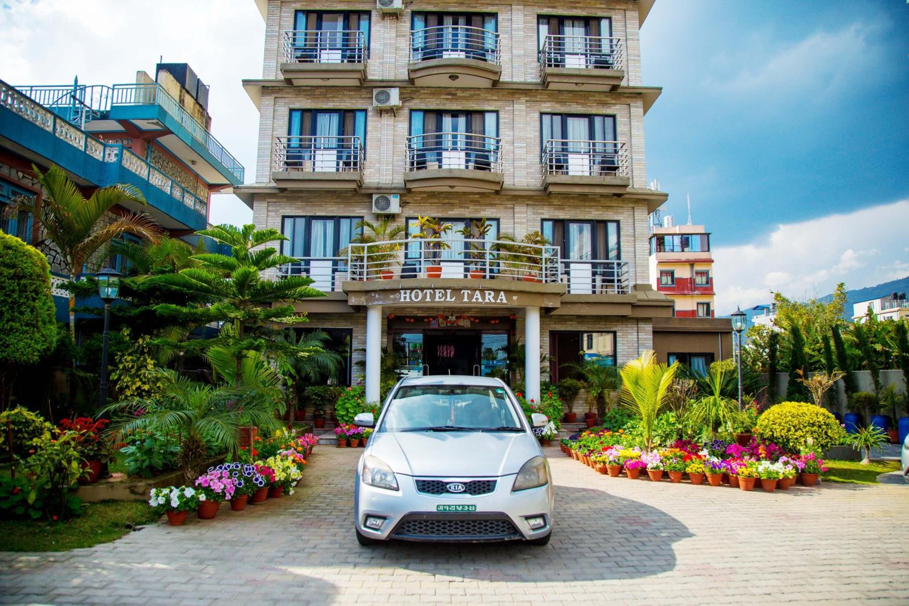 A multi-story hotel with balconies, a car parked in front, and a variety of potted plants.