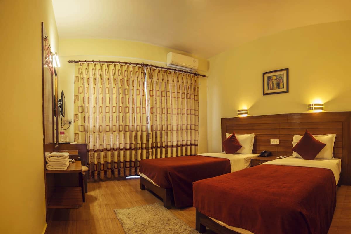 Une chambre d’hôtel avec deux lits simples, une tête de lit en bois, une table de chevet avec une lampe, un miroir et un rideau à motifs géométriques.