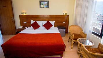 Habitación de hotel con una cama grande, cabecera de madera, una mesita, sillas de mimbre, una ventana con vistas y un cesto de basura.