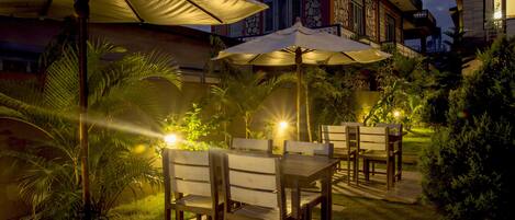 An outdoor patio with tables and chairs, surrounded by plants and a building with a stone facade.