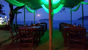 An outdoor dining area with tables and chairs under a green canopy, overlooking a body of water and a distant island. - Cabanas Paraíso (Angra dos Reis)