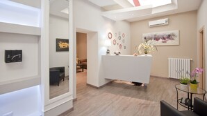 A modern reception area with a white desk, a large mirror, and a ceiling light. There is a wall-mounted air conditioning unit and a framed abstract painting. - Relais Sistina (Rome)