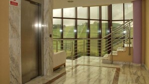 Un pasillo con suelo de mármol, una puerta de ascensor de vidrio, una escalera con barandillas de metal y vistas a un paisaje verde a través de amplias ventanas.