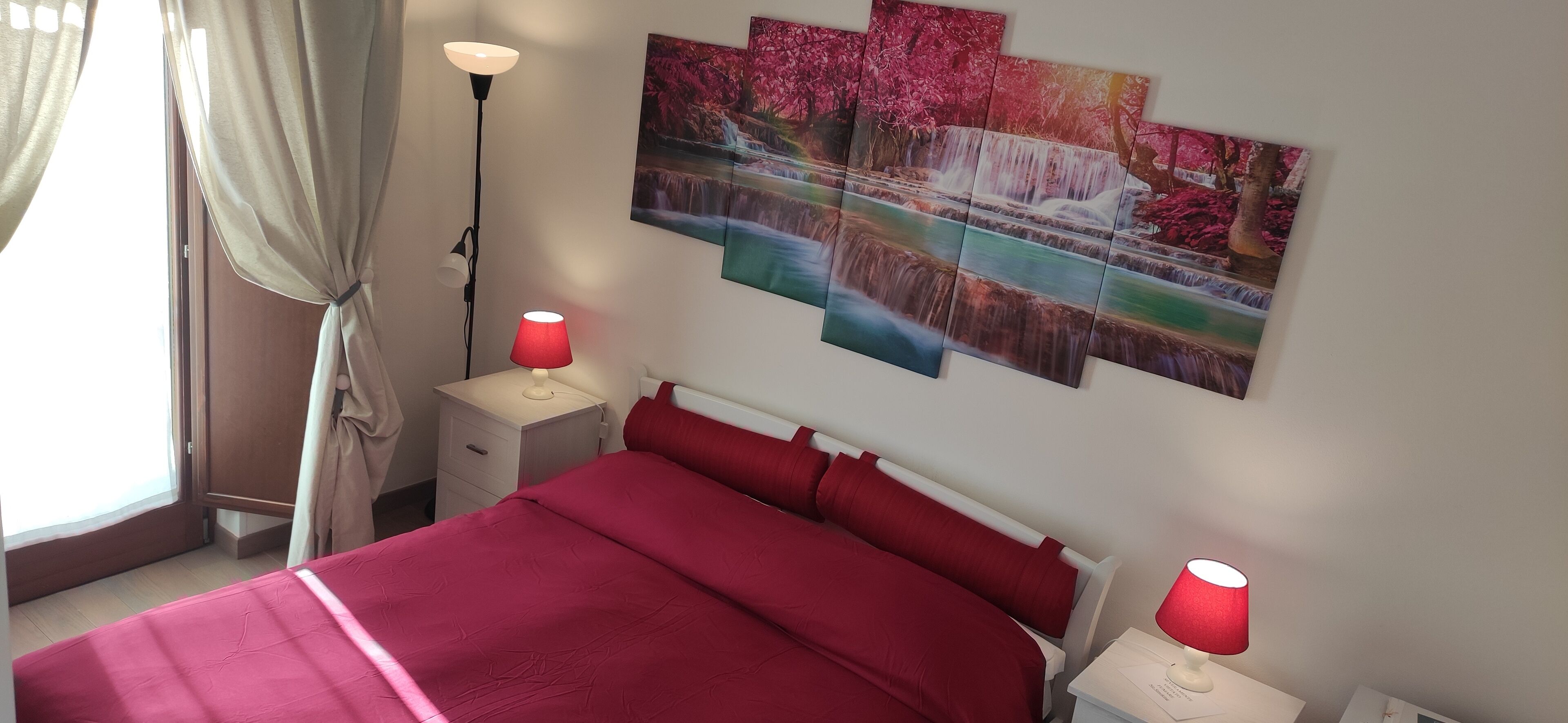Un dormitorio con una colcha roja, dos mesitas de noche con lámparas, una mesita auxiliar y un cuadro de una cascada colgado en la pared.